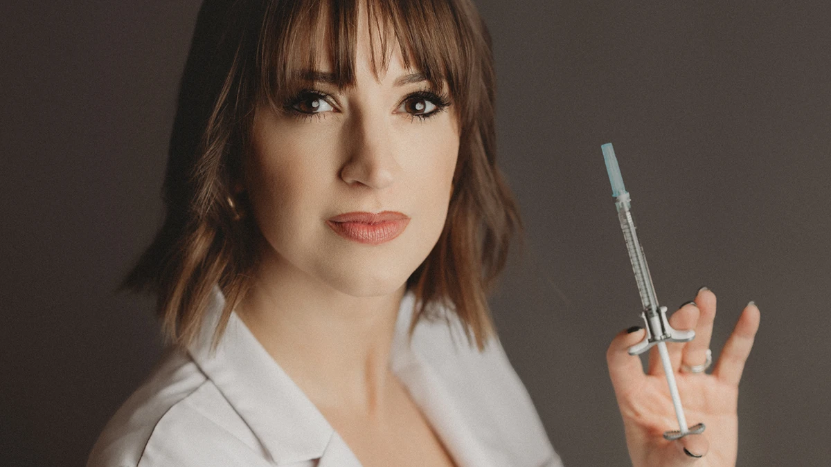 A woman in a white coat holds a syringe, conveying a professional yet calm demeanor against a dark background, suggesting a medical context. A woman in a white coat holds a syringe, conveying a professional yet calm demeanor against a dark background, suggesting a medical context.