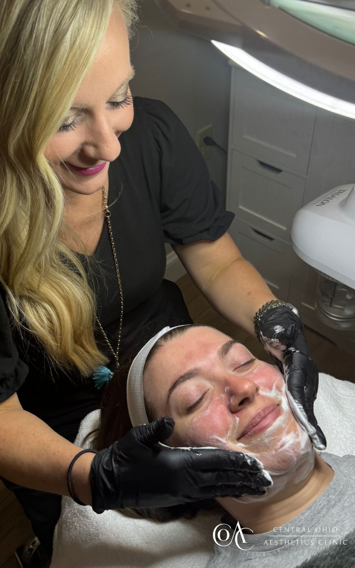 A smiling cosmetologist applies a creamy facial mask to a relaxed client in a spa setting. Warm lighting and calm ambiance convey a soothing atmosphere.