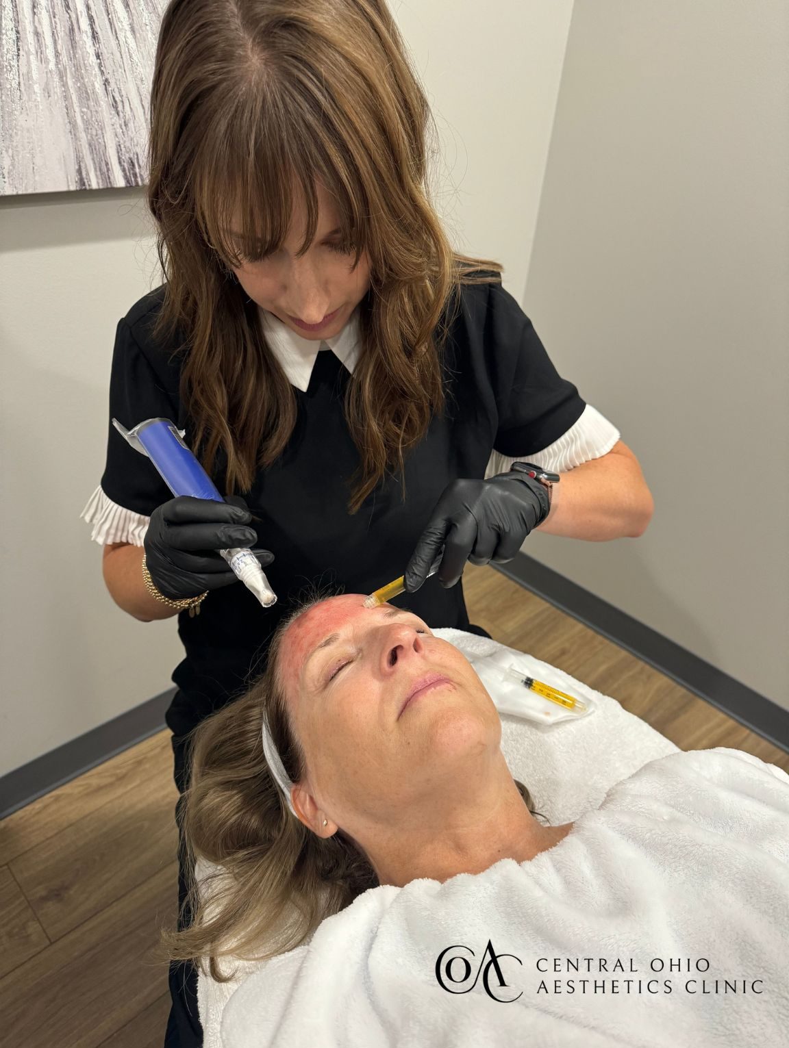 A woman in a clinic receives a facial treatment, lying relaxed on a bed. A practitioner with gloves works carefully on her forehead. The atmosphere is calm and professional.