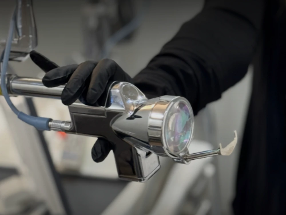 A person wearing black gloves is holding a chrome dental tool with a nozzle and transparent cover, in a clinical setting. The atmosphere is professional.