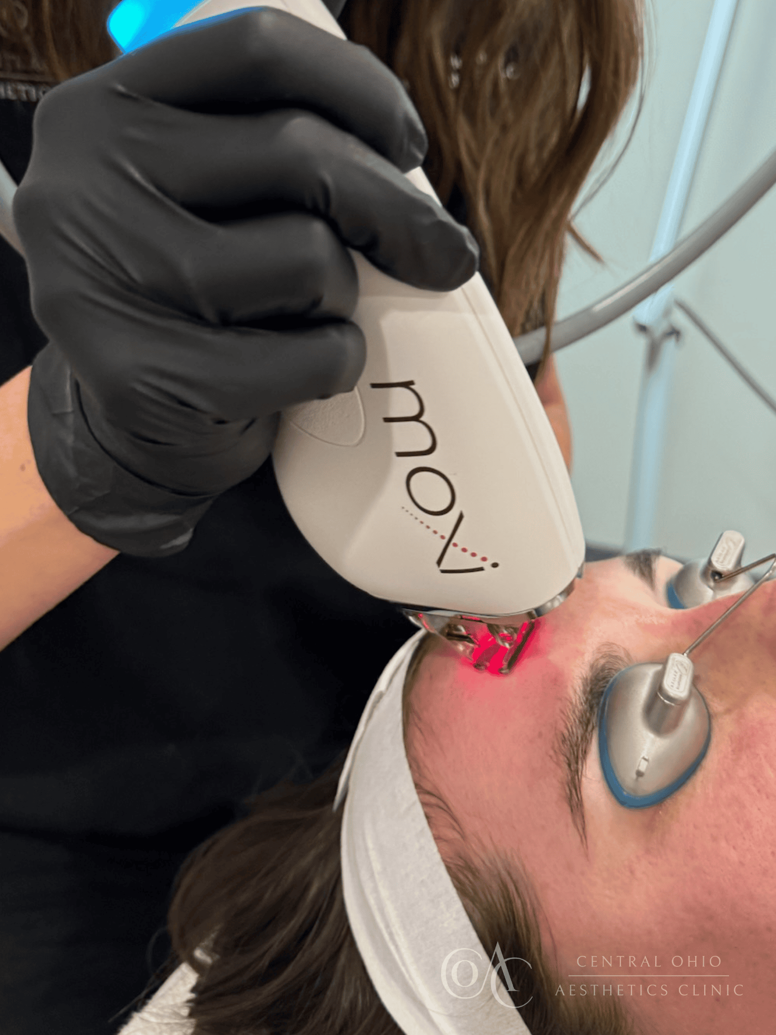 A woman receiving a laser hair treatment at a salon, focused on her hair while a stylist operates the laser device.