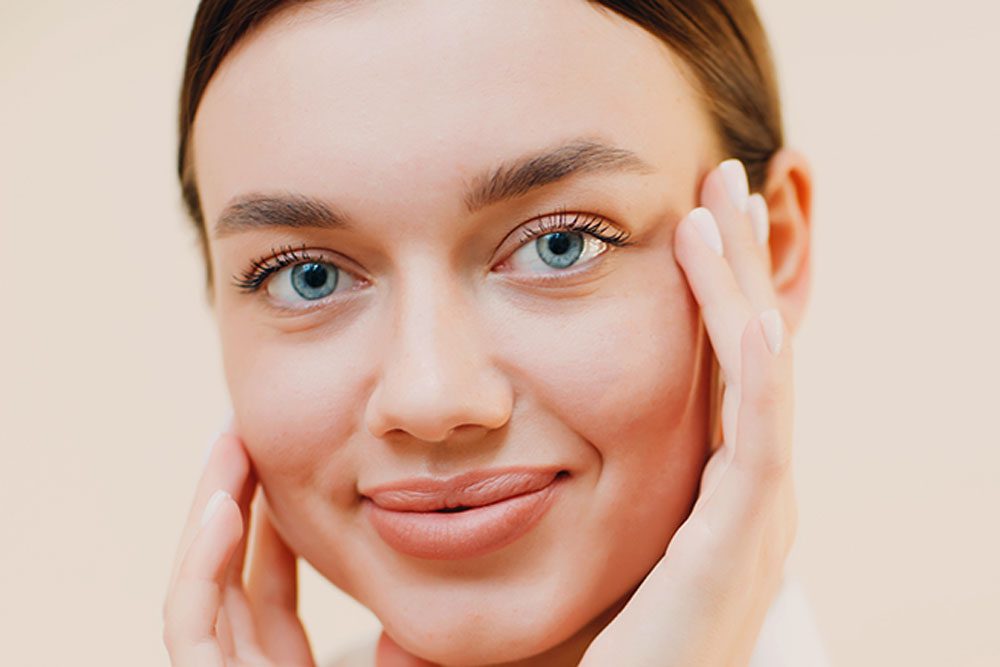 A woman with blue eyes and a gentle smile touches her face with both hands against a soft beige background, conveying a sense of natural beauty and calm.