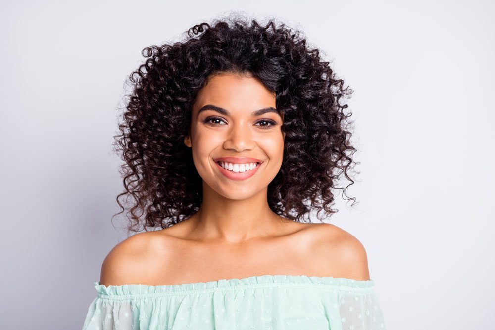 Smiling woman with curly hair in an off-the-shoulder light blue top against a plain background. She exudes a cheerful and confident vibe.