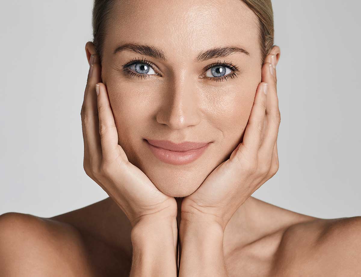 A smiling person with blue eyes and smooth skin gently cups their face with both hands. They have a serene expression. The background is a soft gray.