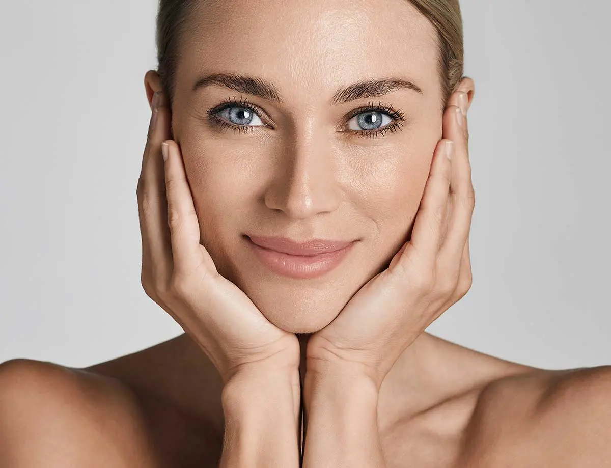 A woman with blue eyes and a calm expression gently holds her face with both hands. Her skin appears smooth and radiant against a neutral background.